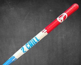 May include: A red, white, and blue baseball bat with the text "2 Chill" in blue. The bat features a peace sign and a dripping paint design. The bat is angled against a dark background.