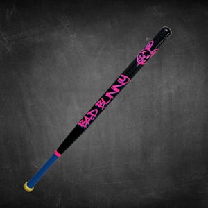 K&ouml;nnte beinhalten: Schwarzer Baseballschl&auml;ger mit pinkfarbenem "BAD BUNNY" Schriftzug und einem Hasen-Grafik. Der Schl&auml;ger hat einen blauen und pinkfarbenen Griff mit gelbem Ende. Der Schl&auml;ger ist vor einem dunklen Hintergrund platziert.