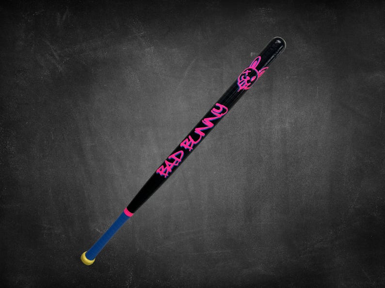 K&ouml;nnte beinhalten: Schwarzer Baseballschl&auml;ger mit pinkfarbenem "BAD BUNNY" Schriftzug und einem Hasen-Grafik. Der Schl&auml;ger hat einen blauen und pinkfarbenen Griff mit gelbem Ende. Der Schl&auml;ger ist vor einem dunklen Hintergrund platziert.
