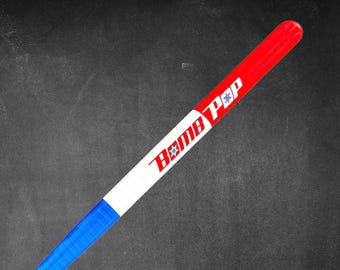 May include: A red, white, and blue Bomb Pop-themed hockey stick. The stick has a red tip, a white center with the words "Bomb Pop" in red, and a blue lower section. The stick is angled against a dark background.