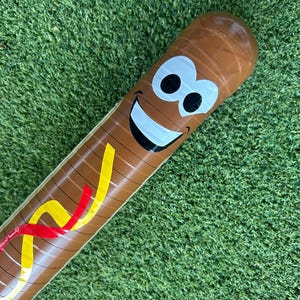 Op de afbeelding: Bruine opblaasbare honkbalknuppel met een lachend gezicht en kleurrijke accenten. De knuppel heeft een cartoon-achtig ontwerp met een witte glimlach, zwarte ogen en gele en rode strepen.