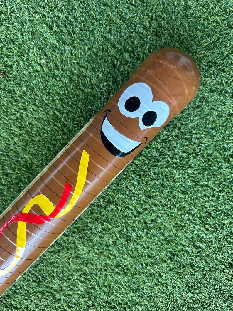 Op de afbeelding: Bruine opblaasbare honkbalknuppel met een lachend gezicht en kleurrijke accenten. De knuppel heeft een cartoon-achtig ontwerp met een witte glimlach, zwarte ogen en gele en rode strepen.