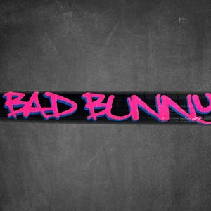 K&ouml;nnte beinhalten: Schwarzes zylindrisches Objekt mit dem Schriftzug "BAD BUNNY" in leuchtend pinkfarbenen Graffiti-Buchstaben, blau umrandet. Der Hintergrund ist eine dunkelgraue, strukturierte Oberfl&auml;che.