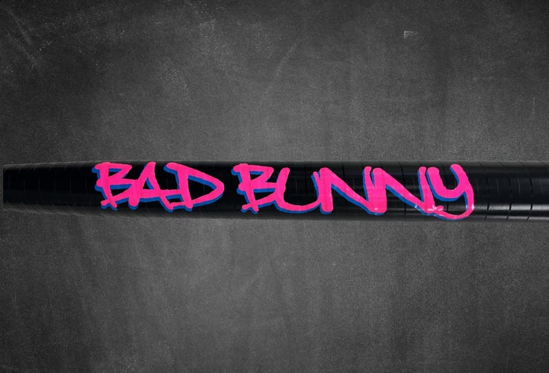 K&ouml;nnte beinhalten: Schwarzes zylindrisches Objekt mit dem Schriftzug "BAD BUNNY" in leuchtend pinkfarbenen Graffiti-Buchstaben, blau umrandet. Der Hintergrund ist eine dunkelgraue, strukturierte Oberfl&auml;che.