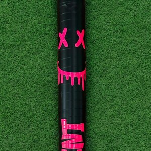 Puede incluir: Objeto cil&iacute;ndrico negro con un gr&aacute;fico rosa ne&oacute;n de una cara sonriente con bordes goteando y dos s&iacute;mbolos 'XX' rosas. Sobre un fondo verde.