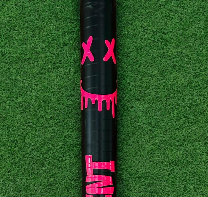 Puede incluir: Objeto cil&iacute;ndrico negro con un gr&aacute;fico rosa ne&oacute;n de una cara sonriente con bordes goteando y dos s&iacute;mbolos 'XX' rosas. Sobre un fondo verde.