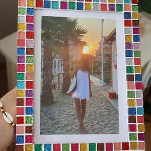 Könnte beinhalten: Ein rechteckiger Bilderrahmen mit einem Rand aus kleinen, quadratischen, glitzernden Fliesen in den Farben Rot, Blau, Grün, Gelb, Pink und Silber. Der Rahmen enthält ein Foto einer Person, die bei Sonnenuntergang an einem Sandstrand steht.