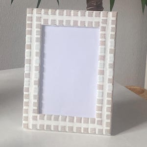Könnte beinhalten: Ein rechteckiger Fotorahmen mit einem Mosaikfliesen-Design. Der Rahmen ist überwiegend weiß mit einem Rastermuster aus kleinen, hellbraunen Fliesen. Der Rahmen steht auf einer weißen Oberfläche.