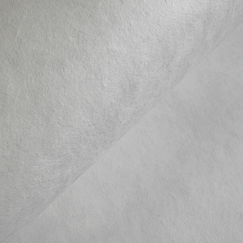 Puede incluir: Primer plano de una tela texturizada de color blanco roto. El material est&aacute; doblado, creando un efecto de capas. La superficie tiene un aspecto ligeramente rugoso y fibroso, con sutiles variaciones tonales. La tela parece suave.