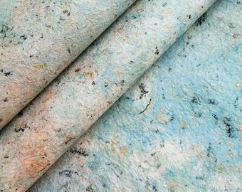 May include: Close-up of a textured, blue, and white paper with flecks of gold and black. The paper is folded in half, showing the texture and color variations.