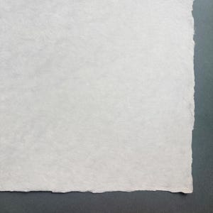 Puede incluir: Primer plano de un trozo de papel blanco hecho a mano con una superficie texturizada y un borde irregular. El papel est&aacute; sobre un fondo gris oscuro.