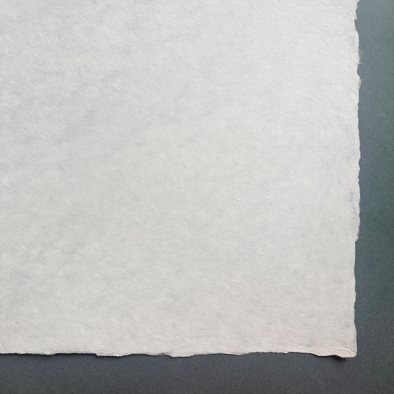 Puede incluir: Primer plano de un trozo de papel blanco hecho a mano con una superficie texturizada y un borde irregular. El papel est&aacute; sobre un fondo gris oscuro.
