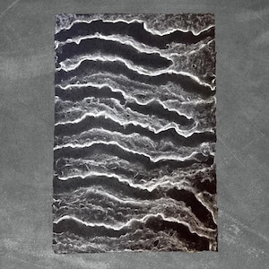 Op de afbeelding: Een rechthoekig kunstwerk met een donkere achtergrond en witte, golvende lijnen. De lijnen creëren een textuur, gelaagd effect, dat lijkt op een topografische kaart of een abstract landschap. Het stuk heeft een handgemaakt, organisch gevoel.