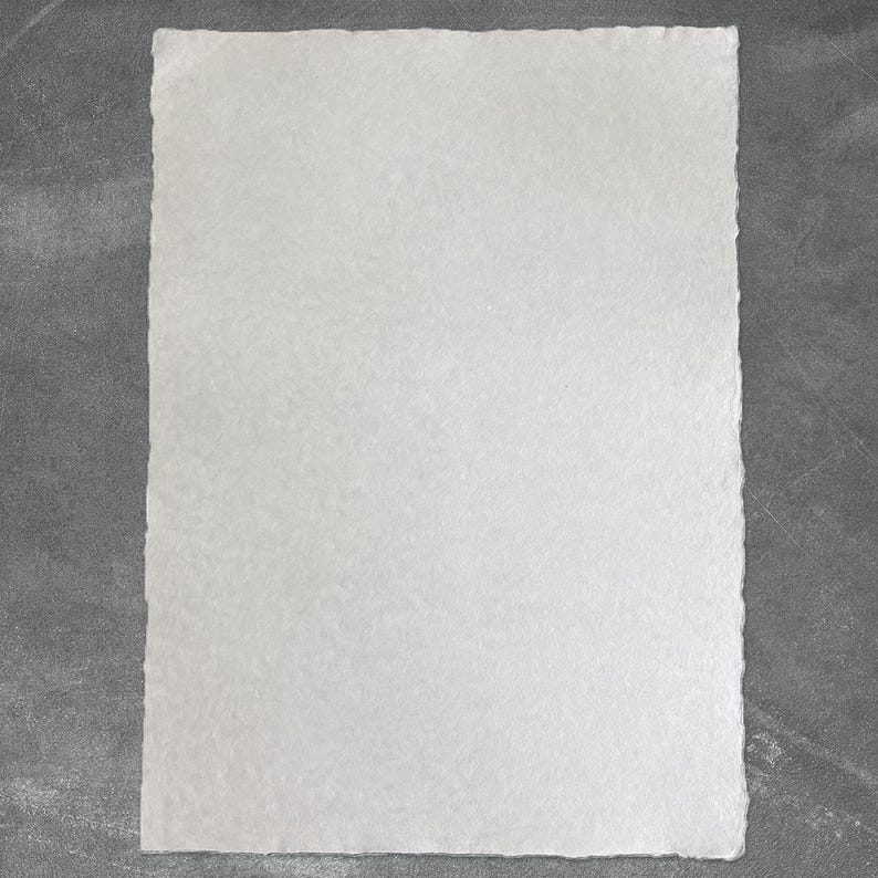 Puede incluir: Una hoja rectangular de papel blanco con una superficie texturizada y bordes irregulares. El papel est&aacute; centrado sobre un fondo gris, creando un contraste. Los bordes del papel son desiguales, a&ntilde;adiendo una cualidad artesanal.