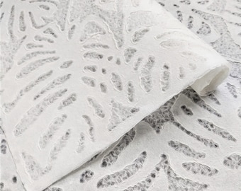 May include: Close-up of a white, textured paper with a cut-out floral design. The paper is partially rolled, revealing the intricate pattern. The design features a repeating pattern of stylized flowers and leaves, creating a delicate and airy aesthetic.