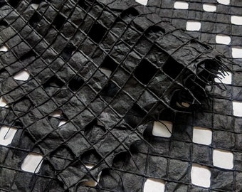 Puede incluir: Papel negro con un patrón de cuadrícula, doblado y superpuesto para crear un efecto texturizado tridimensional. El papel está rasgado y deshilachado en los bordes, lo que le da un aspecto rústico.