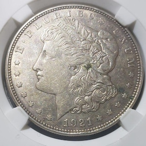 May include: A silver Morgan dollar coin from 1921, displayed in a protective case. The coin features a profile of a woman and the inscriptions "E PLURIBUS UNUM" and "LIBERTY". The coin is round and has a detailed design.