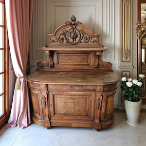 May include: A large, antique wooden cabinet with intricate carvings. The cabinet has a curved front and a decorative top with a central emblem. The wood has a warm, brown tone, and the cabinet is placed in a room with a pink curtain.