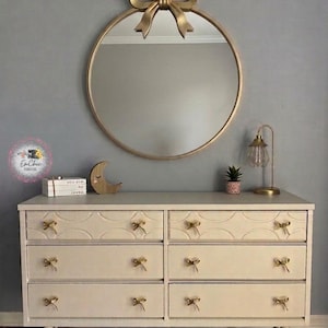 May include: A cream-colored six-drawer dresser with gold bow-shaped drawer pulls. Above the dresser is a round gold mirror with a gold bow accent. A small lamp and potted plant sit on the dresser top.