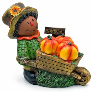 May include: A scarecrow figurine wearing a green plaid shirt and a brown hat with a sunflower, pushing a wooden wheelbarrow filled with three orange pumpkins. A small wooden sign attached to the wheelbarrow features the word welcome in black.