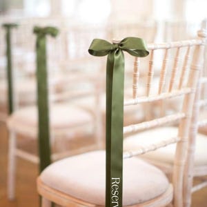 May include: A row of light-colored wooden chairs with cream-colored cushions. Each chair is adorned with a green satin ribbon tied in a bow at the top and a long streamer with the word "Reserved".