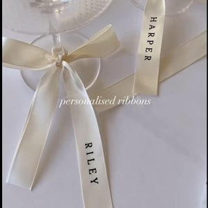 May include: Two champagne glasses with ivory-colored ribbons tied around the stems. The ribbons are printed with names in black text. The image also includes the text "personalised ribbons".