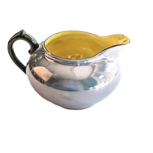 May include: A silver creamer with a yellow interior and a black handle. The creamer is made of ceramic and has a shiny finish.