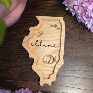 Op de afbeelding: Een lichtgekleurd houten sieradenbakje in de vorm van de staat Illinois met het woord illini in zwarte schrijfletters gegraveerd. Het bakje bevat diverse goudkleurige ringen en kleine oorbellen met een natuurlijke houtnerf.