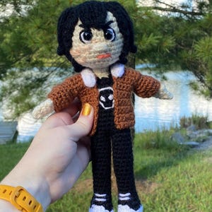 Puede incluir: Muñeca de ganchillo hecha a mano con un personaje de pelo negro, que lleva una chaqueta marrón, pantalones negros y zapatos con ribetes blancos. La muñeca tiene una camiseta negra con un gráfico blanco. La muñeca se sostiene contra un fondo de árboles verdes y un lago.