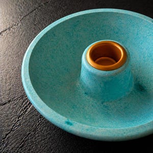 May include: A circular teal candle holder with a speckled texture and a smooth, matte finish. The centre features a raised, cylindrical pillar with a metallic gold interior rim designed to hold a single taper candle securely in place.