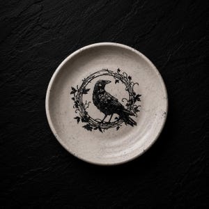 May include: A small, off-white ceramic plate with a black raven illustration encircled by a wreath of leaves. The raven is depicted in a detailed, stylized manner. The plate has a slightly textured surface and is set against a dark background.