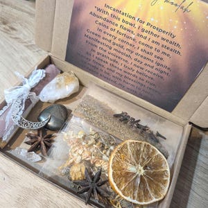 May include: A box containing items for spiritual rituals. Inside are crystals, dried herbs, star anise, a dried orange slice, and a card with the text "Manifesting moolah" and an incantation.