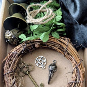 May include: A box containing a rustic wreath, metal charms, a key, and bells. A black satin ribbon and faux greenery are included. The charms include a pentagram and a raven skull. The wreath is made of woven twigs.