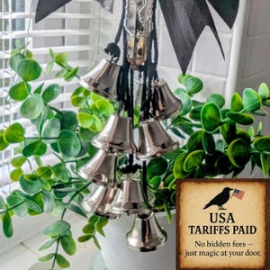 May include: A collection of silver bells suspended from black cords and a black bow, set against a backdrop of green foliage. A sign in the lower right corner reads "USA TARIFFS PAID" with a crow and American flag.