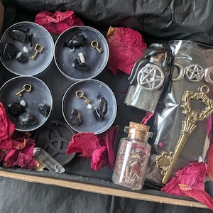 May include: A black box filled with a variety of witchy items, including black tea light candles with black stones and gold keys, a small bottle with a pentagram charm, a bottle of dried herbs, and a gold key charm.