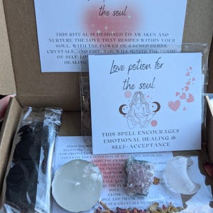 May include: An open cardboard box containing a 'Love Potion for the Soul' kit. The kit includes cards with text, crystals, dried herbs, a candle, and other ritual items. The text on the card says, "This spell encourages emotional healing & self-acceptance."