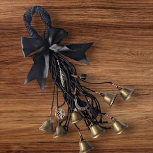 May include: A black rope with a black satin bow and multiple small brass bells hanging from it. The rope is decorated with charms, including a crescent moon, a pentagram, and a green stone.