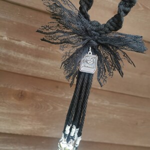 May include: Black and silver charm with a book and pentagram design, with the word "Spells" on the book. The charm is attached to a black cord with a black lace bow and silver bells.