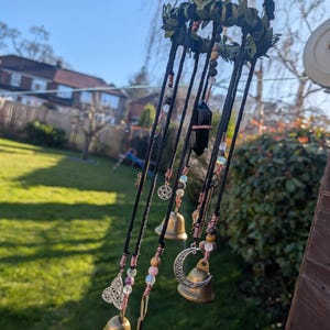 May include: A handcrafted wind chime with a black cord and a decorative wreath. The wind chime features small brass bells, a butterfly charm, a crescent moon charm, and a black crystal. The beads are in various colors, including pink, green, and brown.