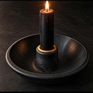 May include: A black pillar candle with a lit flame sits inside a black bowl-shaped candle holder. The candle holder features a central raised circular socket with a thin yellow ring at the base of the candle, providing a contrast to the dark finish.
