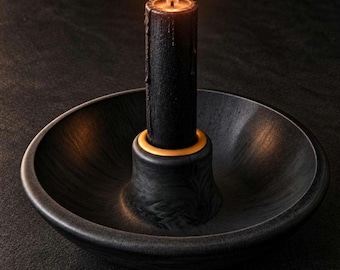 May include: A black pillar candle with a lit flame sits inside a black bowl-shaped candle holder. The candle holder features a central raised circular socket with a thin yellow ring at the base of the candle, providing a contrast to the dark finish.