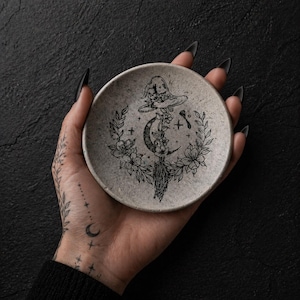 May include: A small, round, speckled gray ceramic dish with a black witch hat, crescent moon, floral, and botanical design. The dish is held in a hand with a tattoo of a crescent moon and stars.