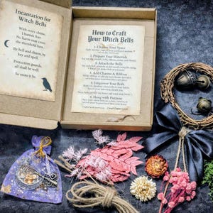 May include: A craft kit in an open cardboard box containing instructions for witch bells, a small purple organza bag with a silver crescent moon and key charm, a bundle of dried pink foliage, twine, a grapevine wreath, and a black satin ribbon.