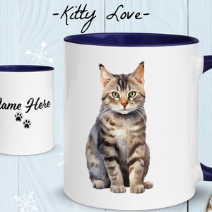 Op de afbeelding: Een witte keramische mok met een blauwe rand en handvat. De mok heeft een aquarel-illustratie van een tabby-kat die zit en naar de camera kijkt. De tekst "Kitty Love" is boven de kat gedrukt en "Name Here" is gedrukt op de andere kant van de mok.
