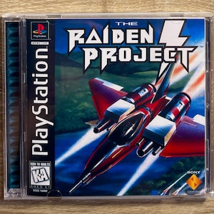 May include: A PlayStation game case for "The Raiden Project." The cover shows a red and black fighter jet over a green landscape. The PlayStation logo and game title are displayed. The game is rated for ages 6+.