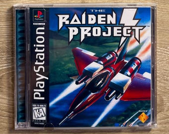 May include: A PlayStation game case for "The Raiden Project." The cover features a red and black fighter jet soaring over a green landscape. The PlayStation logo and game title are prominently displayed.