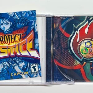 May include: A Dreamcast video game case for "Project Justice." The cover art features a blue and white design with the game title in orange and yellow. The disc inside the case has a red and white flame design.