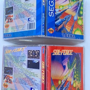 May include: Two Sega CD game cases for the game Sol-Feace. The cases are blue and red with a picture of a spaceship and a dragon on the front. The back of the cases have text describing the game.