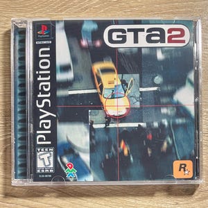 Op de afbeelding: PlayStation-gamecase voor GTA2. De cover toont een bovenaanzicht van een stadsbeeld met een gele taxi in het midden, gericht door een rood vizier. Het PlayStation-logo is links zichtbaar, samen met de ESRB-classificatie.