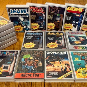 May include: A collection of vintage ColecoVision video game cartridges, featuring titles like "Donkey Kong," "Smurf," and "Space Panic." The game boxes have colorful graphics and text, with a stack of games on the left.